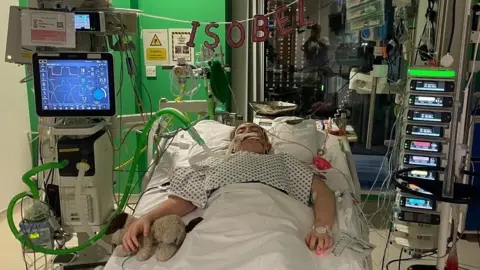Issie Issie is ventilated and lying on a hospital bed in ICU surrounded by medical equipment. She had a teddy bear positioned next to her right hand and is wearing a hospital gown. There is a banner hanging above he bed which reads "Isobel"