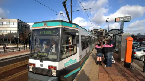 Press Association/John Giles The Metrolink tram link set for Piccadilly on its way around Manchester, stopping at Salford Quays. Passengers are waiting at the stop. It is a sunny day.