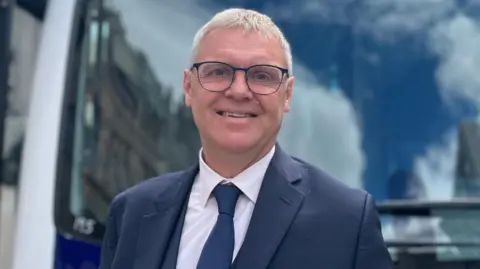 Paul Finch stood in front of the bus and smiling at the camera. He is wearing a suit and tie and glasses. 