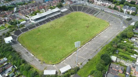 PA Media A grass sports pitch with surrounding grey seats and large floodlights. Around the pitch are residential housing.