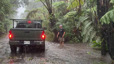 Getty Images A car stops on a rural road as it floods in Volcano Village on Hawaii Island. A man stands next to the car as rain pelts down on a leaf-lined road