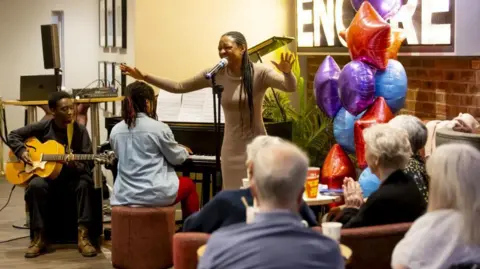 Scott Bird Beverley Knight singing in front of a microphone, her eyes shut. She is wearing a beige tight fitting long-sleeved dress and her hair is long and in braids. She is accompanied by a woman on a piano and a man on guitar to her right. The backs of five people's heads, who all have white or greying hair, can be seen watching from tables. They are in a cafe and there are blue, purple and red helium balloons by a brick wall on the far right.