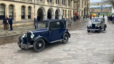Two Austin 7 cars driving past an entrance to the City Hall 