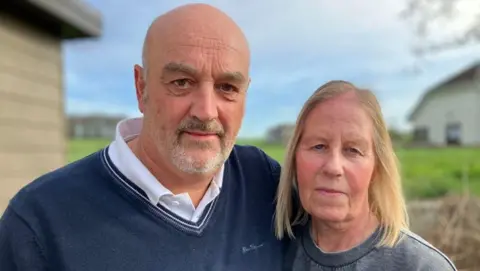 Mark and Sandra Thompson are standing outside in front of a static caravan. Mark is wearing a dark blue sweater over a white collared shirt, and Sandra is wearing a gray sweatshirt. The background shows green grass, another building, and a partly cloudy sky.