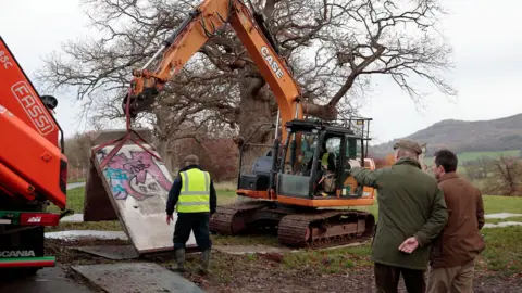 Eastnor Castle An orange crane with large tracks holds up a section of concrete while a man in a high viz jacket supervises. Two other men in green and brown coats watch on in the foreground. 