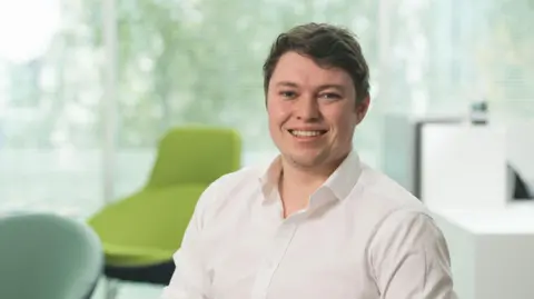 Leigh Day Ryan is seen sitting down on a chair wearing a white shirt, with the sleeves folded up to his elbow. Ryan has blackish-brown hair and is sitting in a room with many green chairs in the background