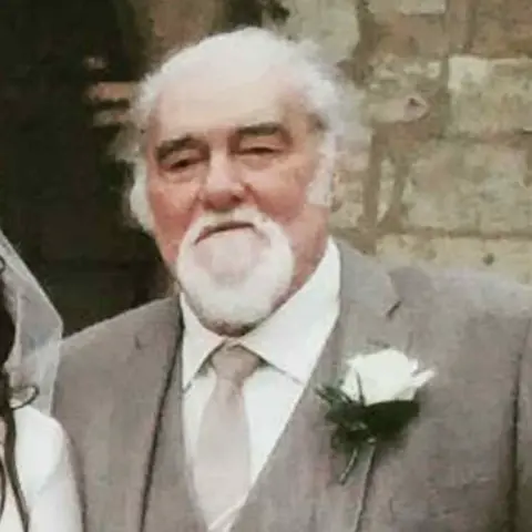 Jordan Wharton Robert has grey hair and a grey beard. He is wearing a grey suit and tie over a white shirt and there is a white flower pinned to his left lapel. He is standing in front of a church.