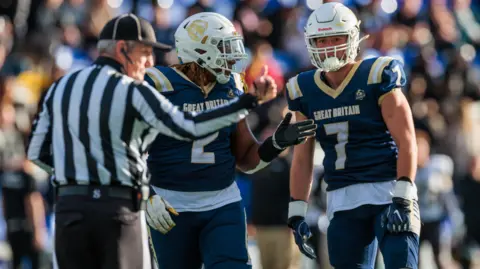 Ben Rogers / BAFA Two British American football players wearing white and navy blue kit and helmets. There is a referee wearing black and red giving the two players an instruction.