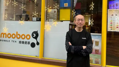 Henry Luk A bald man with glasses in a long-sleeve top standing outside a yellow bubble tea shop.