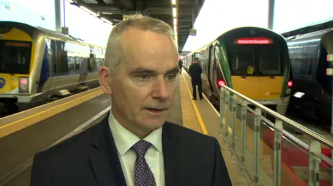 Chris Conway - he wears a black suit, white shirt and colourful tie. he stands in front of the train