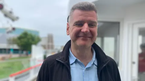 Phil Cunliffe/BBC A middle-aged man stands outside wearing a light blue button-up shirt and and a black jumper. He is smiling. 