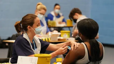 PA Media Students receive vaccines in the sports hall at the University of Kent campus in Canterbury. Nurses in masks and white aprons and NHS lanyards are sitting at tables to give the jabs. there are boxes and yellow buckets on the tables and cotton wool swabs.