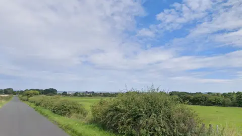 Google A green field site with a rural road alongside 