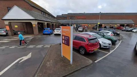 A Google Maps image of a car park in front of a Sainsbury's supermarket. There are dozens of cars parked and a handful of pedestrians walking. 