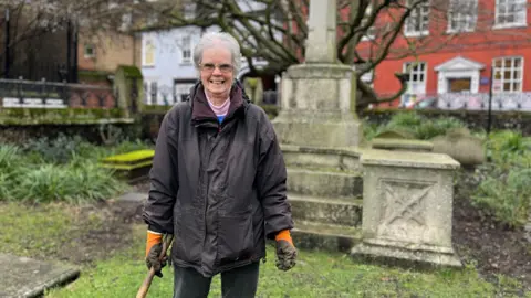 Jon Wright/BBC June Molloy standing with a garden trowel in front of a stone memorial in the church's grounds, smiling at the camera. She has white hair, glasses and is wearing a wax jacket over various layers of clothing.