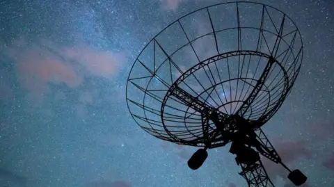 A radar disc pointed a dark night sky with an array of stars and clouds, tinged in purple, visible. The radar itself is in silhouette. 