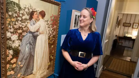 Charles Dickens Museum Lucinda Hawkesley, a middle aged woman with light brown hair, wears a blue velvet dress as she stands in the Charles Dickens Museum and smiles as she looks at a Victorian painting of two of her ancestors, Dickens' daughters.