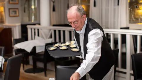 Empire Hotel David, who is wearing a white shirt, black tie and black waistcoat, placing small silver dishes of butter on a table in a large dining room.