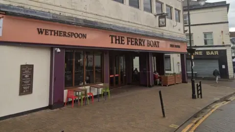 Google Streetview The Ferry Boat in Church Street, Runcorn, situated on a corner plot with an orange sign and black writing, and purple frontage. There are two tables with four chairs outside.