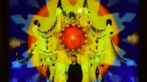 Yellow, red and blue lights are projected onto a huge sculpture of a face. A man dressed in black stands in front of the sculpture.