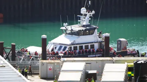 A ship is docked at a port. It is full of people. Most of them are wearing orange life jackets. 