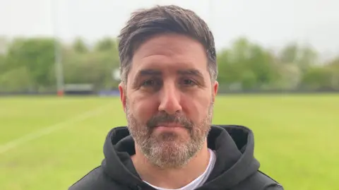 James Burridge/BBC A man with brown hair and short beard wearing a brown hooded top standing on a football pitch. 