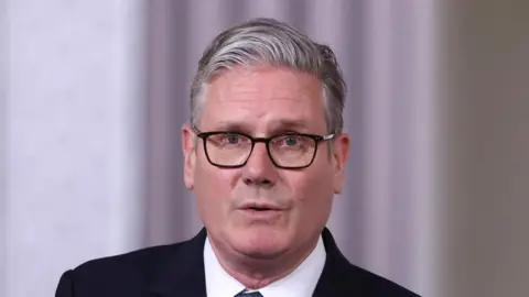 Sir Keir Starmer is looking towards the camera. He has short, grey hair and is wearing a dark suit jacket, a white shirt and a tie.