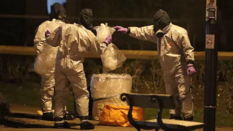 PA Media Three people in full protective, white clothing with purple gloves and a black head covering. They are holding plastic bags above a pop up bin. They are outside, standing near a bench and there is an orange bag on the floor.