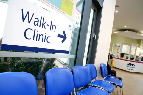 Reception area of the first GP walk-in clinic at Wester Hailes Healthy Living Centre in Edinburgh - six blue chairs are below a sign reading Walk-In Clinic, with an arrow pointing to a reception desk marked with the NHS Lothian logo. 