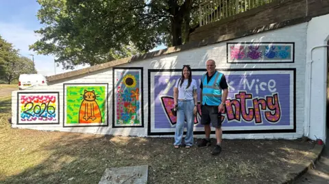 West Northamptonshire Council Two people by a newly painted underpass, with art painted on it. There is a woman in a white top, blue jeans and long dark hair and a man standing next to her with shorts on and a blue high-vis jacket. 