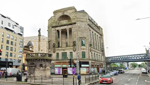 A large building. The ground floor has various posters and vacant signs covering it. Large windows are visible on the other higher floors. 