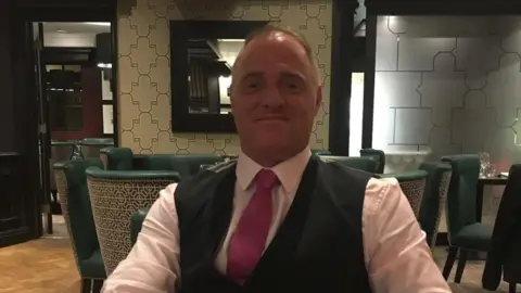 A man in his fifties with a receding hairline dressed smartly in a waistcoat and tie sitting at a table in a restaurant smiling at the camera.