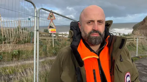 Rosstam Asadi, from Redcar and Cleveland Council stands wearing a green jacket over the top of a fluorescent orange vest. He is posing in front of the cliff face on Cowbar Bank, which now has a protective fencing around it due to erosion. 