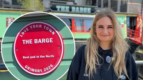 Woman with long blonde hair smiles to camera. She is wearing a dark, navy jacket with her name 'Lily' embroidered into it. In the background The Barge, a bright mint green boat with red detailing around the glass windows can be seen. There is also a close-up picture of the red plaque which reads: 'Love Your Town, The Barge, Best pub in the North, Grimsby 2026'.