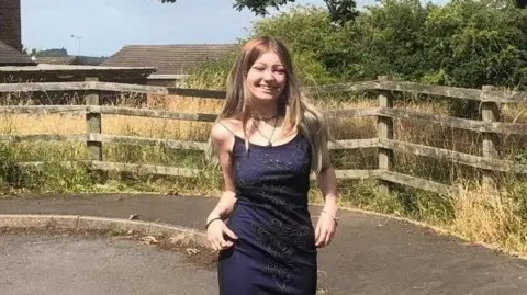 Morgan Dorsett wearing a dark blue dress with silver sequins forming a pattern on the front. She is smiling at the camera, and has long blonde hair with darker roots. She is wearing black eyeliner, a choker necklace and bracelets on each wrist. Behind her there is a curving pavement and a wooden fence, overlooking a patch of dry grass and a large green bush on a sunny day.