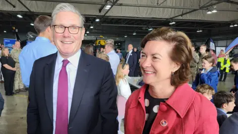 A man with grey hair, glasses, a navy suit, blue shirt and a red tie - Sir Keir Starmer - smiles while standing next to a woman with brown hair, wearing a navy polka-dot dress and red jacket - Eluned Morgan.
