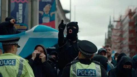 Masked protesters and police officers face each other in a city street during a public demonstration. Several people wear dark clothing and face coverings, while two police officers in high-visibility jackets stand in the foreground. A large blue-and-white vehicle or display is visible behind the crowd, with city buildings and scaffolding in the background.