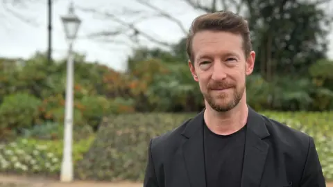 Douglas is wearing a black jacket and dark top stands on a gravel path in a park, with trees, shrubs, and a lamppost in the background.