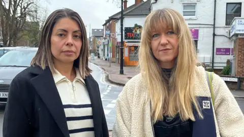 BBC Two women stand next to each other in front of the camera, with a busy high street behind them. The woman on the left has shoulder-length black hair and is wearing a cream and blue striped top and navy jacket, the woman on the right has long blonde hair and is wearing a cream-colored fleece