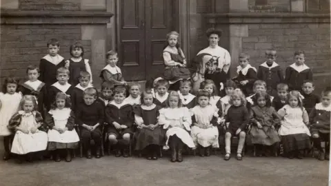Saltaire World Heritage Education Association A black and white photo of a large group of children in Victorian era clothes. They are posing in school photo style with a woman in the middle who looks like a teacher.