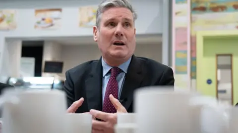 PA Media Sir Keir Starmer wearing a light blue shirt, maroon tie with dots and a dark blazer. His mouth is open, caught speaking mid-sentence, and he is gesturing with his hands
