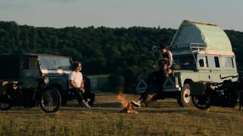 On the Roam/HBO Max Momoa and others sat on the hood of two vintage Land Rovers around vintage Harley-Davidson motorcycles. They are parked around a fire in a field.