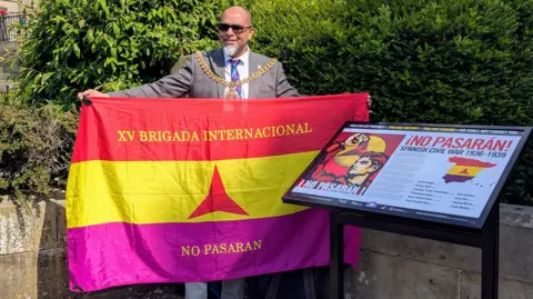 Shah Hussein holds an International Brigade flag, which has red, yellow and crimson horizontal stripes, along with the words 'No pasaran'. The memorial, which includes the words 'Spanish Civil War 1936 - 1939' can be seen to his left.