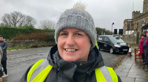 A middle-aged woman smiles as she stands on a village street on a grey and damp winter day. She is wearing a grey bobble hat with a white fluffy pompom, a black jacket and a yellow hi-vis vest.