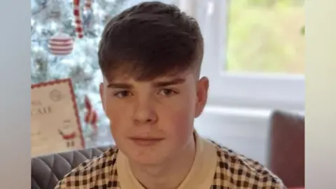 A headshot of a boy with short brown hair, combed in a side shed. He is expressionless and appears to have been photographed in a living room with a Christmas tree in the background. He is wearing a brown and white checked polo shirt.