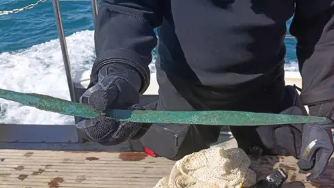 A person wearing a dark diving suit and gloves is standing on a boat and holding up a long, corroded green metal sword-like object, possibly recovered from the seabed. The boat's wooden deck is wet, with a mesh bag and diving equipment nearby, while blue sea water and the boat's railing can be seen in the background.