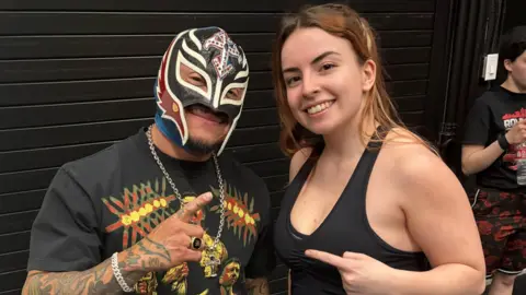 Natasha Swann Rey Mysterio and Natasha Swann point towards each other while standing in a gym. He is wearing a black mask and black T-shirt. Natasha is wearing black work-out gear.