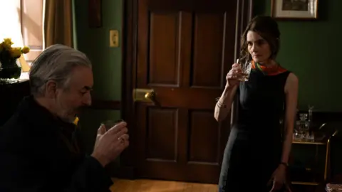 Graeme Hunter Two people hold drinks in a warmly lit room with dark green walls, polished wooden doors and traditional furnishings.
One person sits in the foreground while the other stands nearby, creating a sense of distance and tension within the scene.
Soft natural light from a window illuminates the interior, highlighting the rich wood tones and intimate atmosphere.