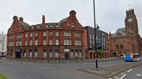 The Majestic Hotel in its current form. It is a red-brick, four-storey building with large windows on the corner of a road. Its gables on either side look like rounded triangles. There are short black safety railings in front of it. In the background is a church clock tower and a bus is going past.