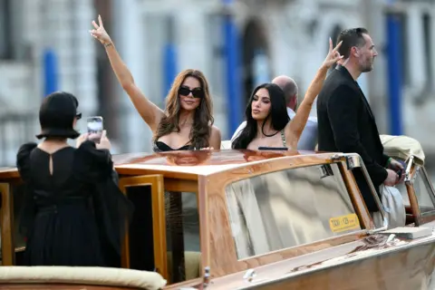 Getty Images Kris Jenner, Khloe Kardashian and Kim Kardashian in Venice, Italy, for the wedding of Jeff Bezos and Lauren Sanchez. They are gesturing peace signs on a taxi boat while their mother Kris Jenner photographs them. 